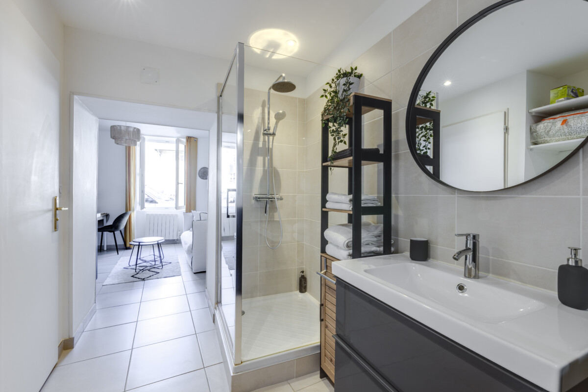 Salle de bain Herriot 5 Brive – Douche vitrée, meuble vasque gris, miroir rond et accès direct au séjour lumineux