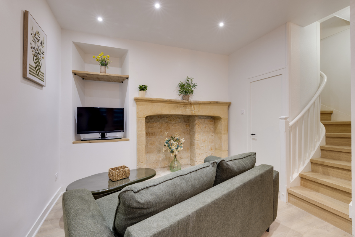 Salon lumineux et élégant avec canapé convertible confortable, cheminée en pierre, télévision et escalier menant à l’étage dans un appartement de charme à Sarlat-la-Canéda.